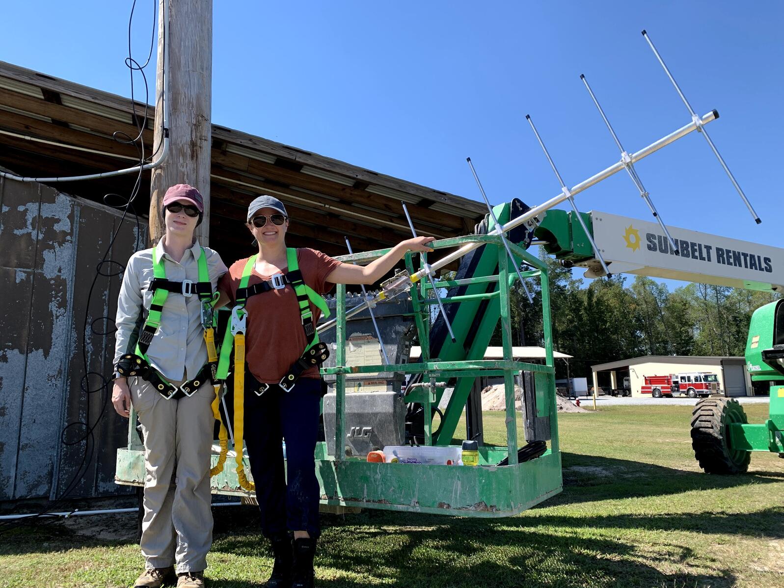Jen and Heather wear safety harnesses and stand next to a boom lift and antenna at a Motus Installation site. 