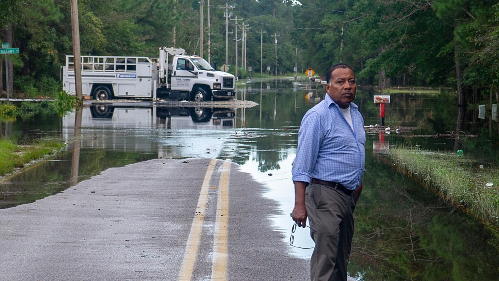 A man stands in front of a flooded road with emergency vehicles in the background