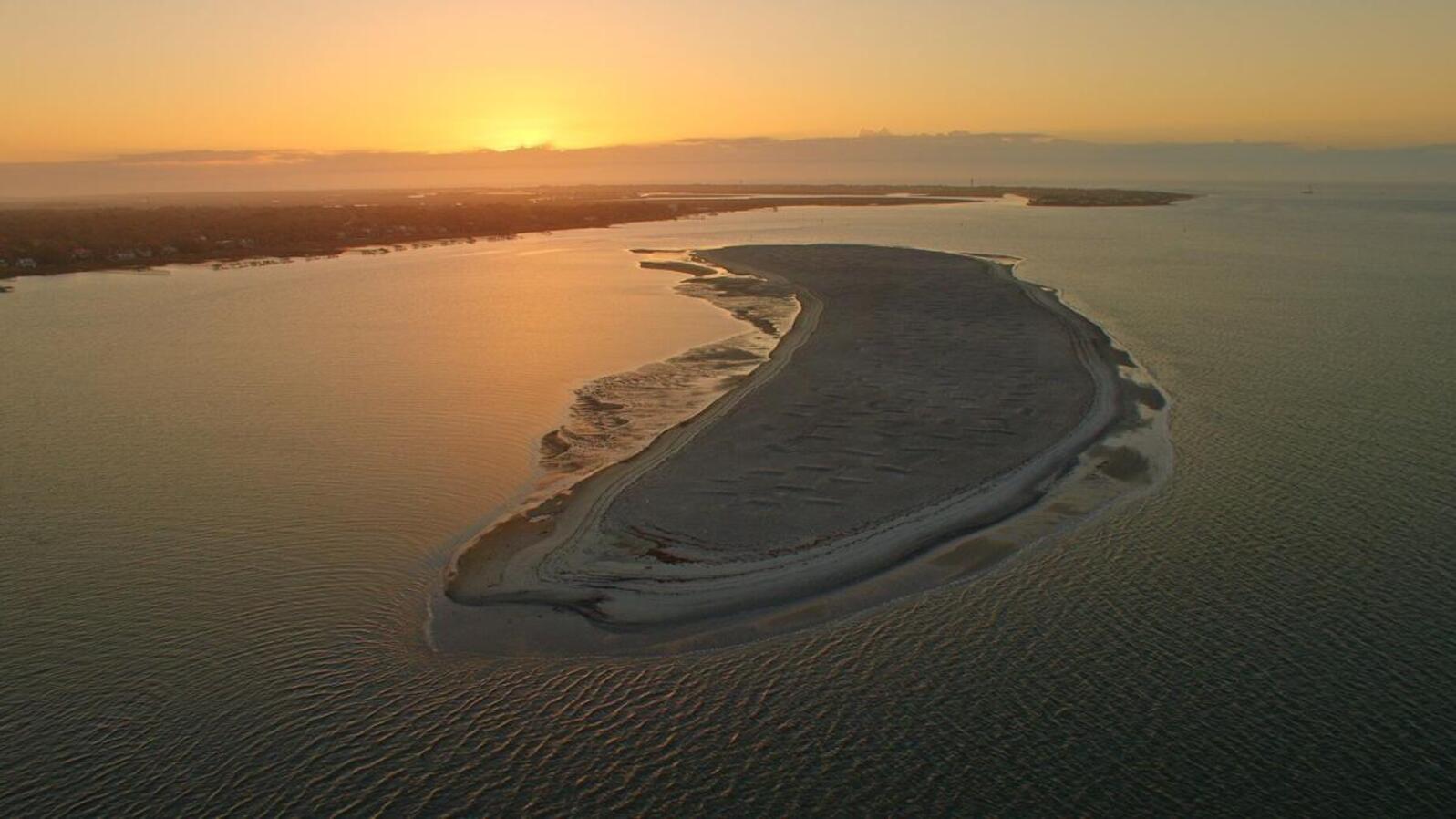 An aerial view of a large sand island at sunset, bathed in an orange glow with calm waters of Charleston harbor lapping around it. 