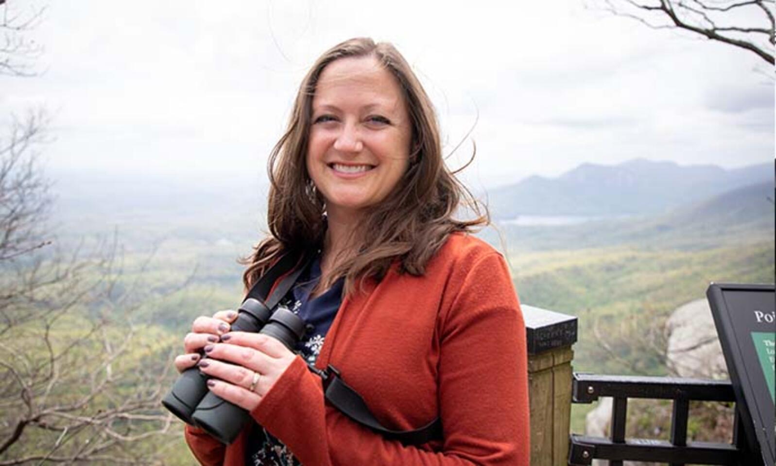 Jen Tyrrel Smiles holding binoculars at the Caesar's Head State Park overlook with a vast landscape view of green mountains and a cloudy sky