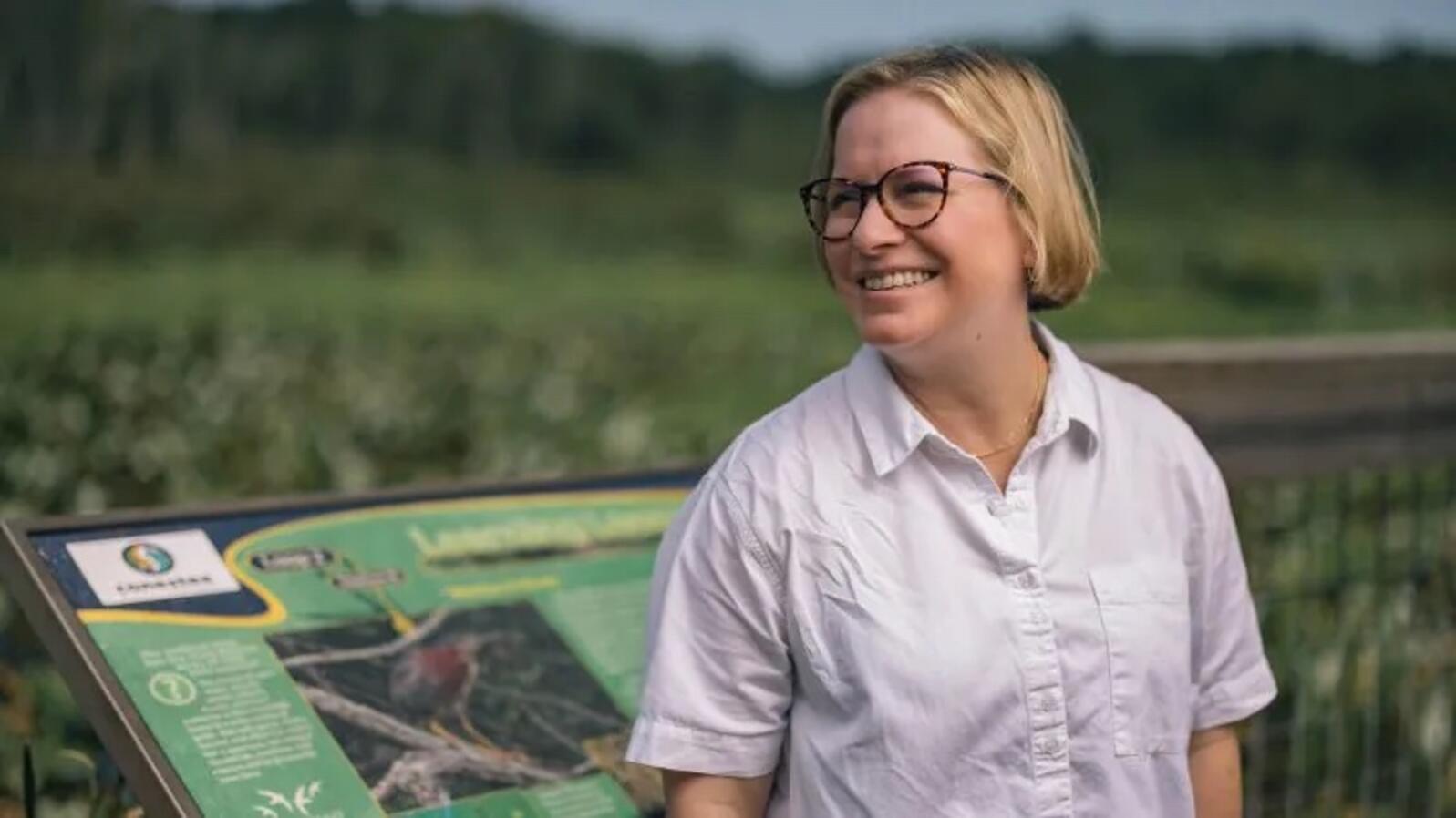 Rebecca Haynes smiles over her shoulder in front of a blurry green background of wetlands and distant forests with an interpretive sign at her elbow.